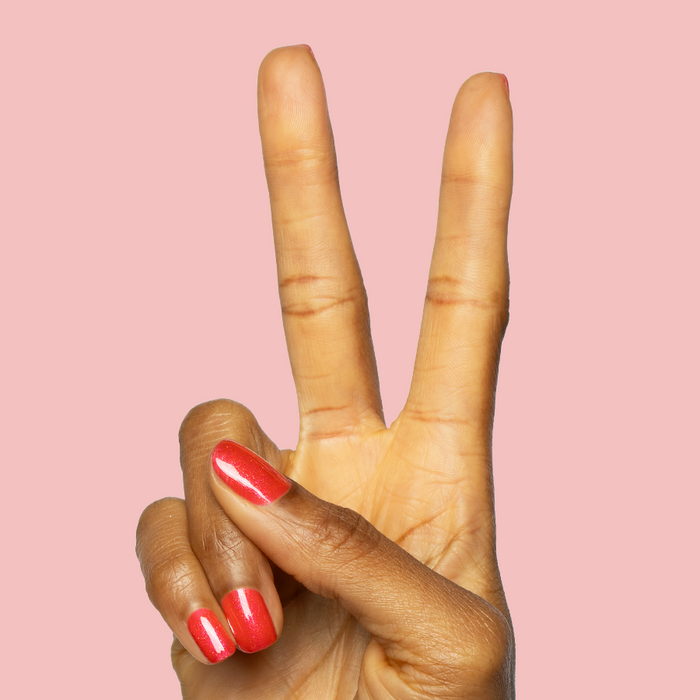 Hand showing peace sign with nails painted bright pinky-red shimmer using Rodeo Queen - Gel polish on pink background